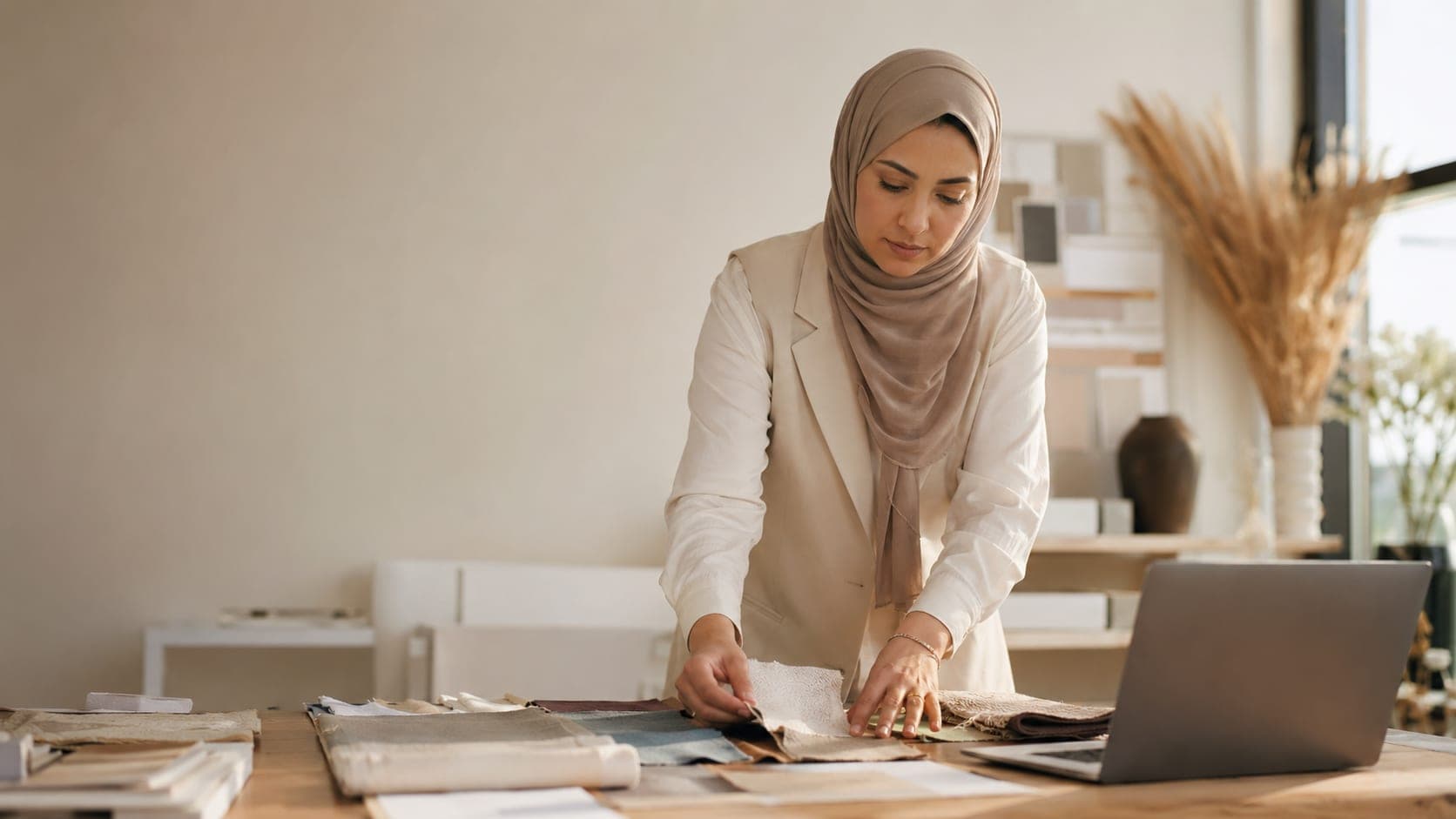 Een vrouw met hoofddoek legt stofstalen op een werktafel naast een laptop.