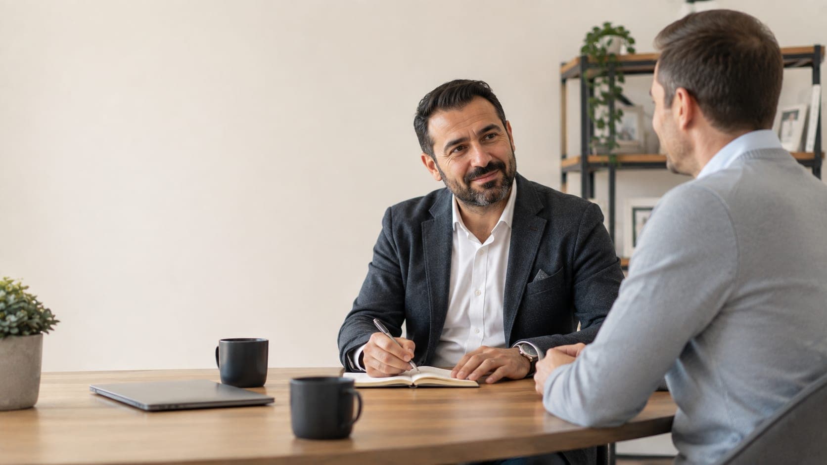 Twee mannen zitten aan tafel in gesprek; een van hen schrijft in een notitieboek.